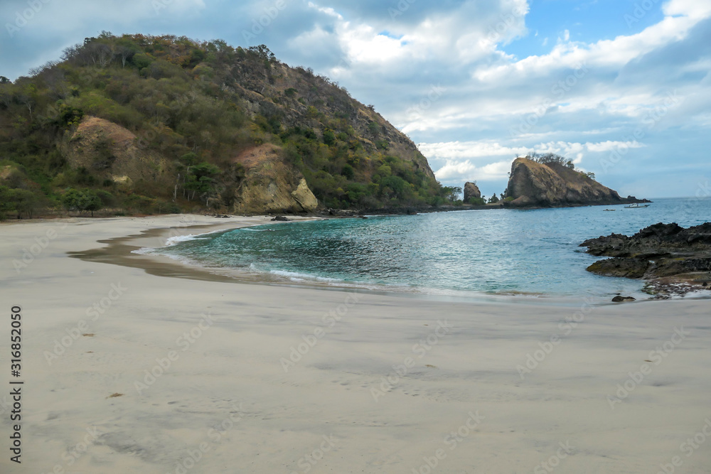 A panoramic view of idyllic Koka Beach. Hidden gem of Flores, Indonesia. Beach is gently washed by waves. Steep hills going straight into the sea. Soft clouds on the sky. Serenity and calmness