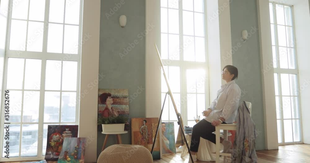 Workflow of paintress is drawing a picture on canvas near the easel, side view. Paintress is painting picture sitting in modern art studio near big windows, view from afar and camera zooms in. Workflow of paintress is drawing a picture on canvas near the easel, side view. Paintress is painting picture sitting in modern art studio near big windows, view from afar and camera zooms in.