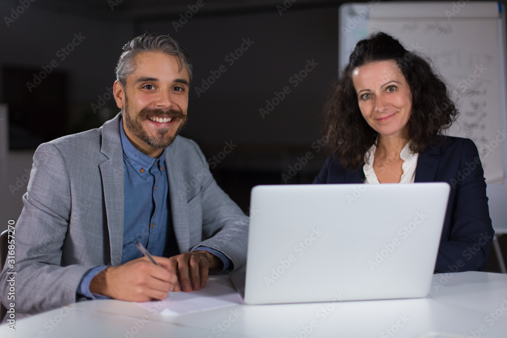 Smiling colleagues sitting at table and looking at camera. Cheerful office managers sitting at table with laptop. Teamwork, working late concept Smiling colleagues sitting at table and looking at camera. Cheerful office managers sitting at table with laptop. Teamwork, working late concept