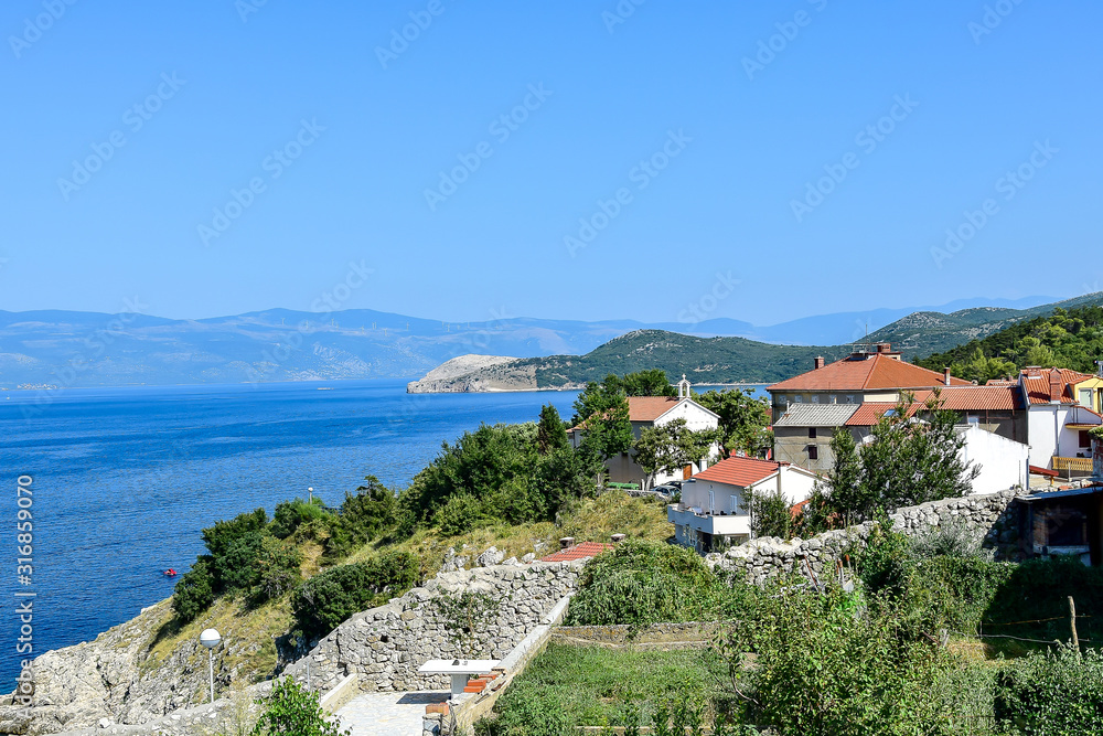 Vrbnik / Krk, Croatia - City, white and beige houses with orange roofs, green trees, blue sky with clouds, mountains and the sea in the background, in the summer afternoon.