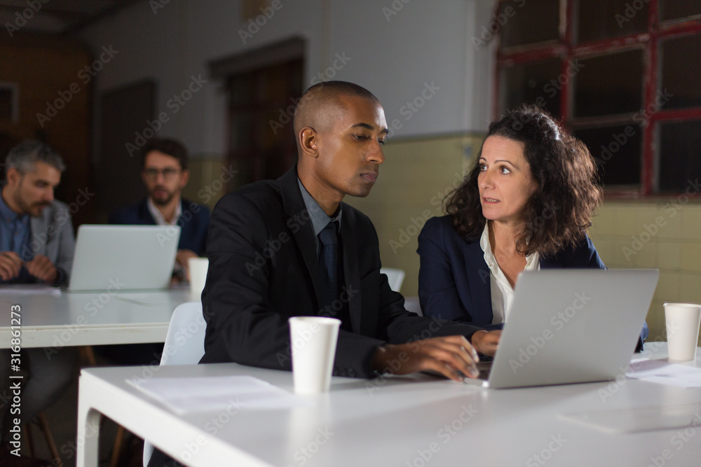 Confident employees talking while working with laptop. Focused business partners discussing business questions in office. Business, working late concept Confident employees talking while working with laptop. Focused business partners discussing business questions in office. Business, working late concept