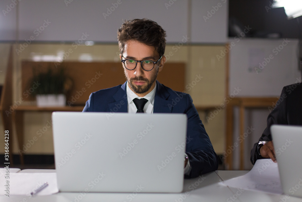 Front view of focused man in eyeglasses looking at laptop. Concentrated handsome manager sitting at table and working with laptop. Business, working late concept Front view of focused man in eyeglasses looking at laptop. Concentrated handsome manager sitting at table and working with laptop. Business, working late concept
