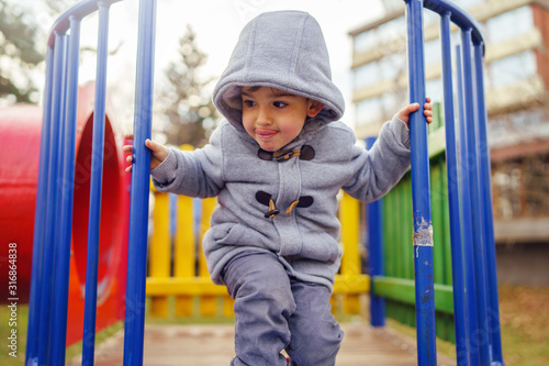 Portrait of small little caucasian boy kid wearing winter coat walking standing on the slide playground in the park with hood on his head in winter day smiling