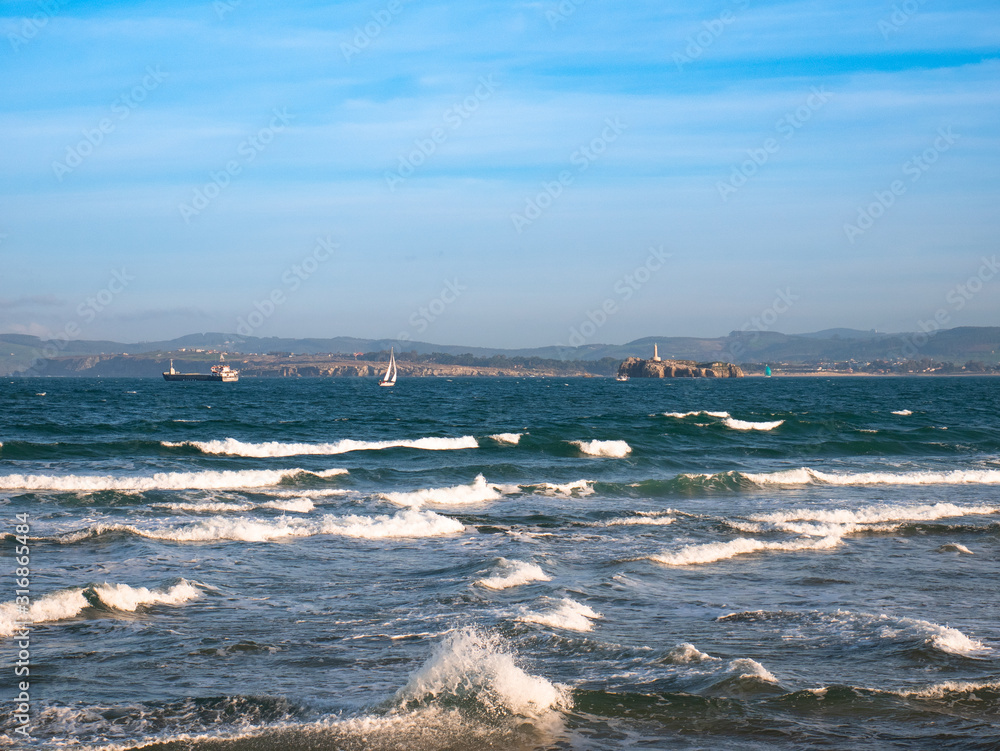 Obraz premium An oil tanker and a sailboat sail along the Cantabrian Sea near the bay of Santander with the lighthouse and the Cantabrian coast in the background