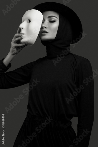 high fashion portrait of elegant woman in black dress and hat, holding theatre white mask. Studio shoot. Concept of magic actor face 