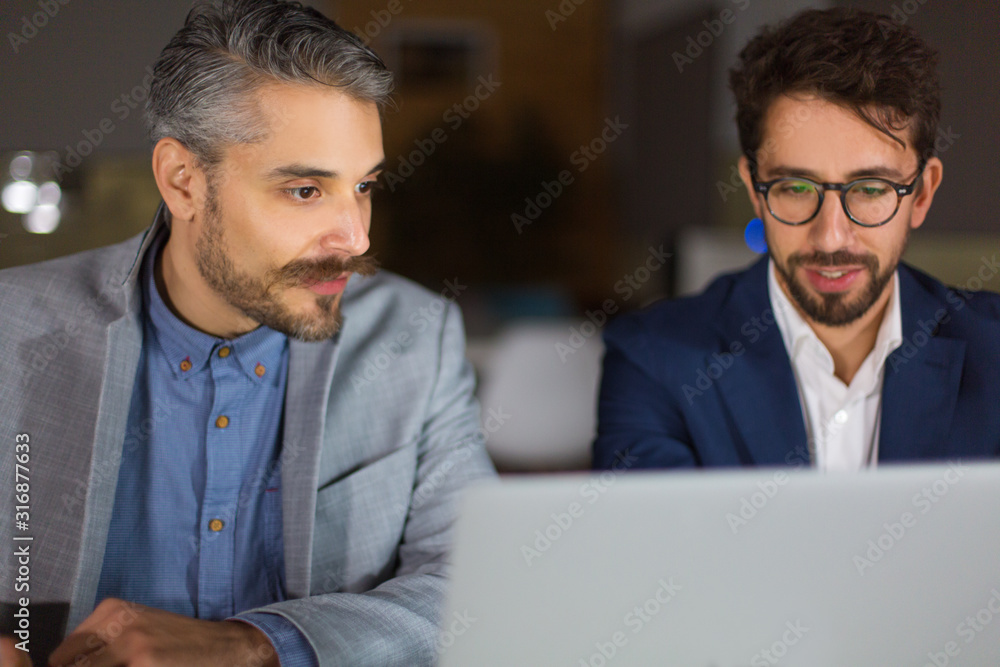 Obraz premium Two handsome businessmen looking at laptop. Front view of two business partners working with laptop. Business, working late concept