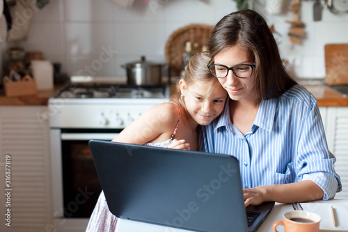 Mother and daughter using laptop and Internet. Freelancer workplace in cozy kitchen. Woman and child girl together. Concept of female business, working mom, freelance, home office. Lifestyle moment.