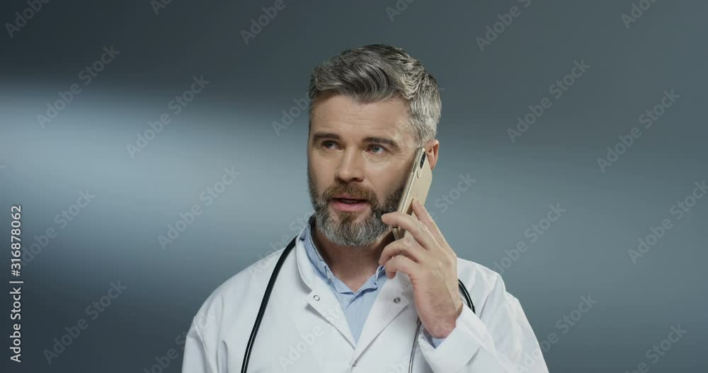 Caucasian male physician speaking on the mobile phone and doing consultation by connection on the gray wall background. Caucasian male physician speaking on the mobile phone and doing consultation by connection on the gray wall background.
