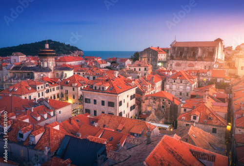 Aerial view of houses with red roofs at sunset in Dubrovnik, Croatia. Top view of beautiful architecture in european old city. City lights, historical centre, buildings and blue sky at dusk in summer