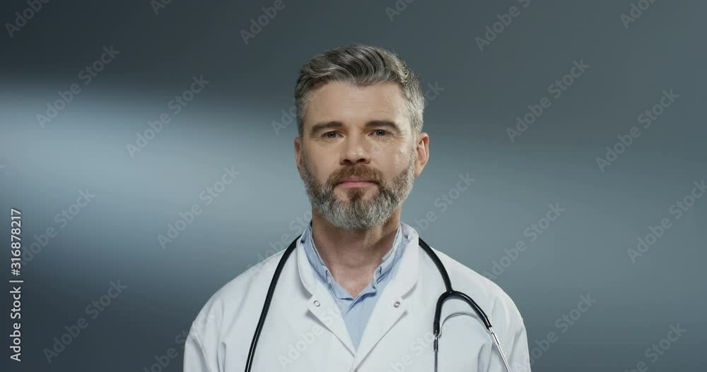 Portrait shot of the good looking Caucasian man physician in white gown and with stethoscope on his neck rising face up and smiling to the camera. Close up. Portrait shot of the good looking Caucasian man physician in white gown and with stethoscope on his neck rising face up and smiling to the camera. Close up.