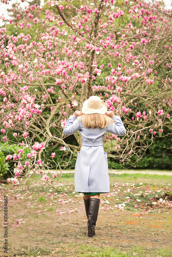 a young, beautiful woman, in a blue coat and a light hat, walks in the Park in the spring near a flowering Magnolia tree, on a Sunny day