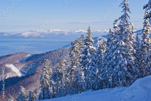 A view from the heights of the ski resort of the mountain peaks overgrown with fir trees and an unfrozen lake in winter.