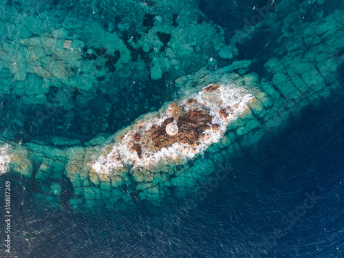 Above aerial view of lighthouse on a tiny island in the middle of the sea. Colorful water color. Clear water allows you to see the topography of the sea floor. Sardinia island in Mediterranean sea.
