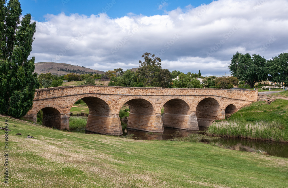 Richmond, Tasmania, Australia - December 13, 2009: Landscape and entire brown stone historic bridge over coal river under blue cloudscape with green lawn up front and dark green trees around.