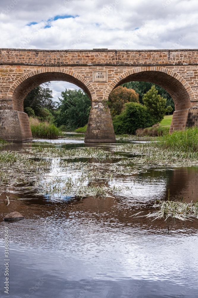 Richmond, Tasmania, Australia - December 13, 2009: Aong water line closeup portrait shot of part of brown stone historic bridge over coal river reflected in water with reed. Some blue cloudscape