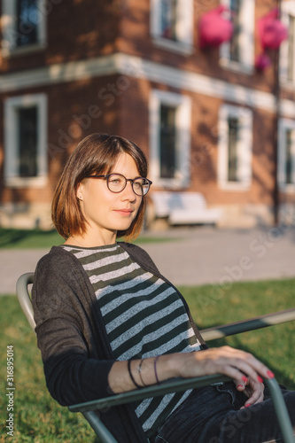 Close up sunny portrait of charming woman in glasses and striped t-shirt. Pretty girl sitting in outdoor chair on green grass. Brick building on background. Recreation in park. Summertime concept.