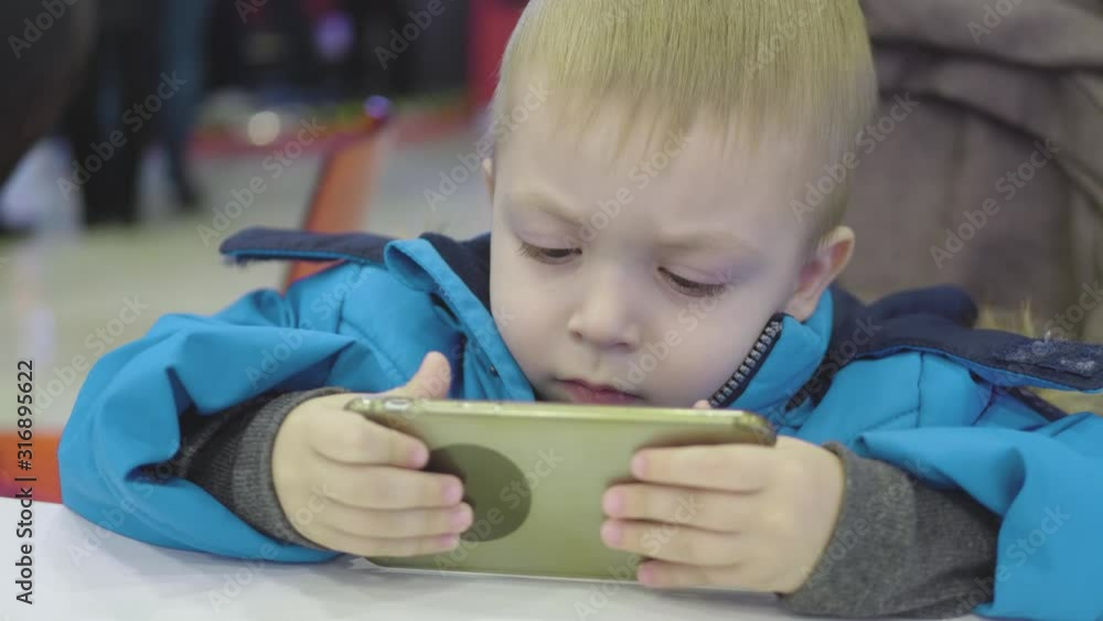 Cute little boy sitting in the cafe watching cartoons on phone with parent. Cute little boy sitting in the cafe watching cartoons on phone with parent.