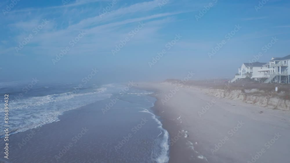 Moving fast over the beach shoreline. The waves are crashing on the beach as it moves down the coast. Dunes and houses line the shore. Moving fast over the beach shoreline. The waves are crashing on the beach as it moves down the coast. Dunes and houses line the shore.