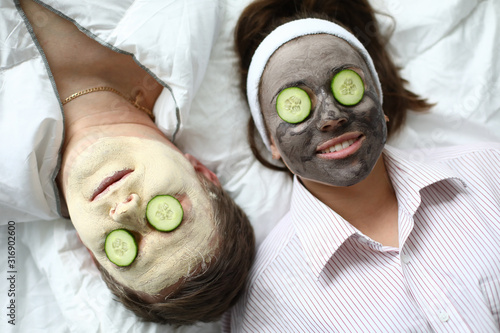 Top-view of man and woman making spa procedures applying wetting facial masks and fresh cucumber on eyes. Couple laying on bed. Wellness and skincare concept