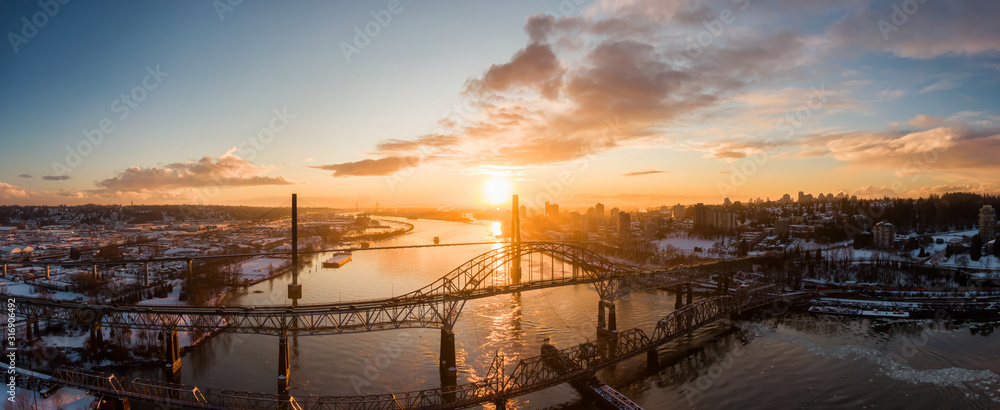 Fototapeta premium Aerial Panoramic View of the Modern City, Bridge and Fraser River during a colorful Sunset. Taken in New Westminster, Vancouver, British Columbia, Canada.