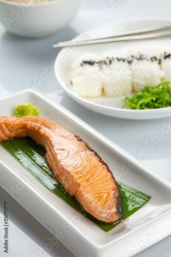 Grilled salmon with soy sauce with Japanese rice topped with black sesame seeds in a white plate on a white table