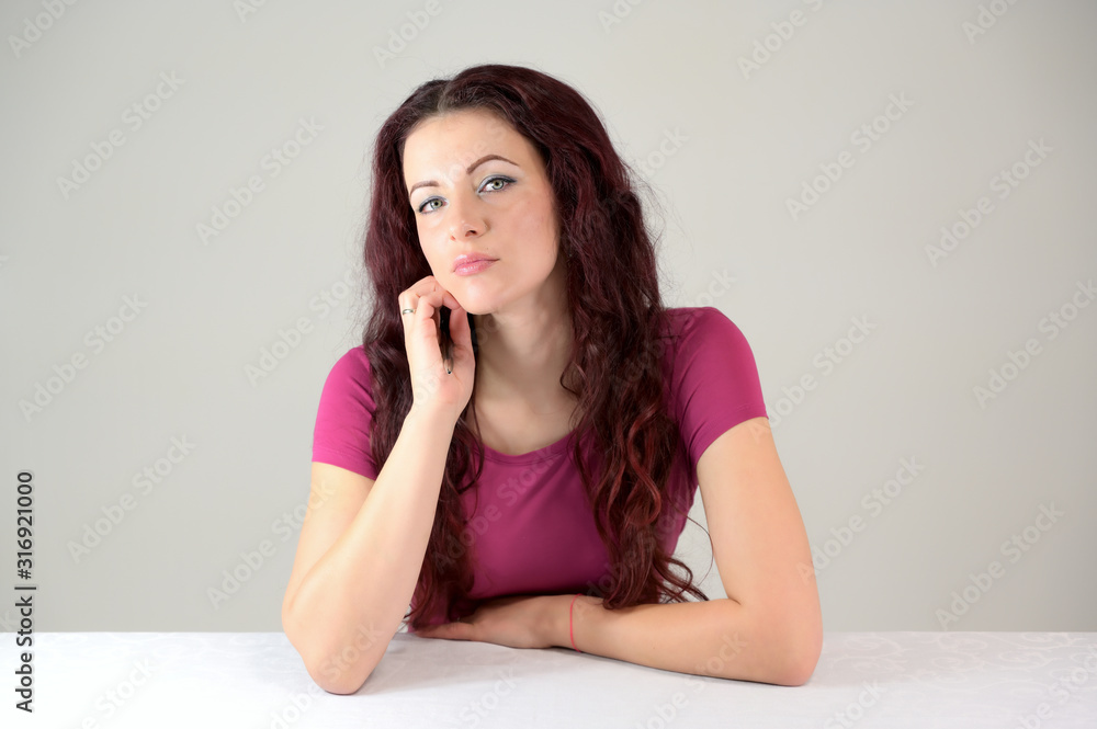 The model smiles and enjoys posing and talking. Portrait of a pretty slender brunette woman in a pink dress and with brown hair sitting at a table on a white background in the studio. The model smiles and enjoys posing and talking. Portrait of a pretty slender brunette woman in a pink dress and with brown hair sitting at a table on a white background in the studio.
