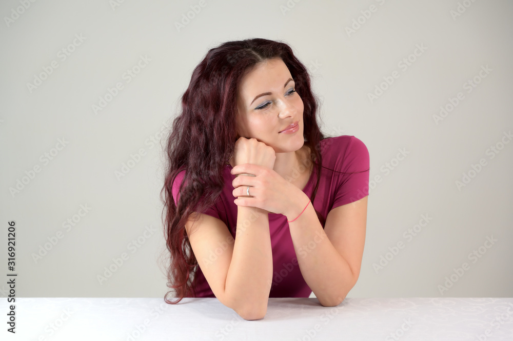 Model is smiling and talking on the phone. Portrait of a pretty slender brunette woman in a pink dress and with brown hair sitting at a table on a white background in the studio. Model is smiling and talking on the phone. Portrait of a pretty slender brunette woman in a pink dress and with brown hair sitting at a table on a white background in the studio.