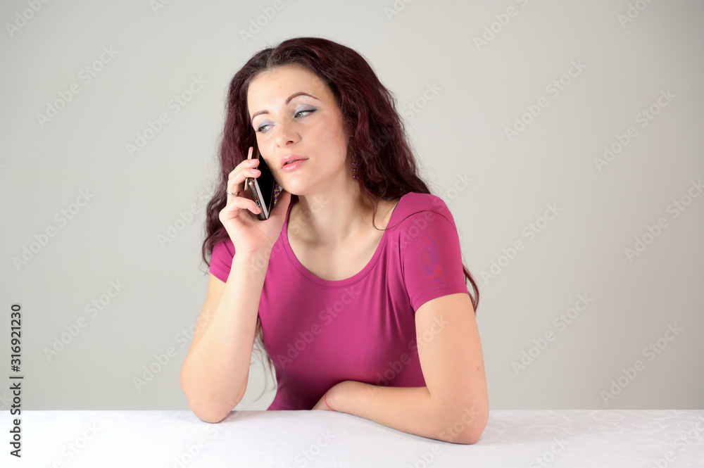 Model is smiling and talking on the phone. Portrait of a pretty slender brunette woman in a pink dress and with brown hair sitting at a table on a white background in the studio. Model is smiling and talking on the phone. Portrait of a pretty slender brunette woman in a pink dress and with brown hair sitting at a table on a white background in the studio.