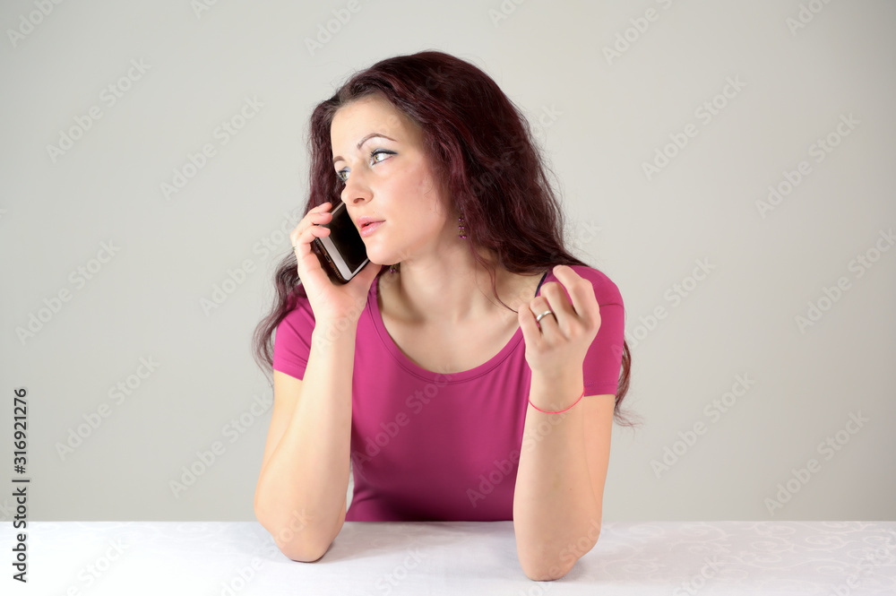 Model is smiling and talking on the phone. Portrait of a pretty slender brunette woman in a pink dress and with brown hair sitting at a table on a white background in the studio. Model is smiling and talking on the phone. Portrait of a pretty slender brunette woman in a pink dress and with brown hair sitting at a table on a white background in the studio.