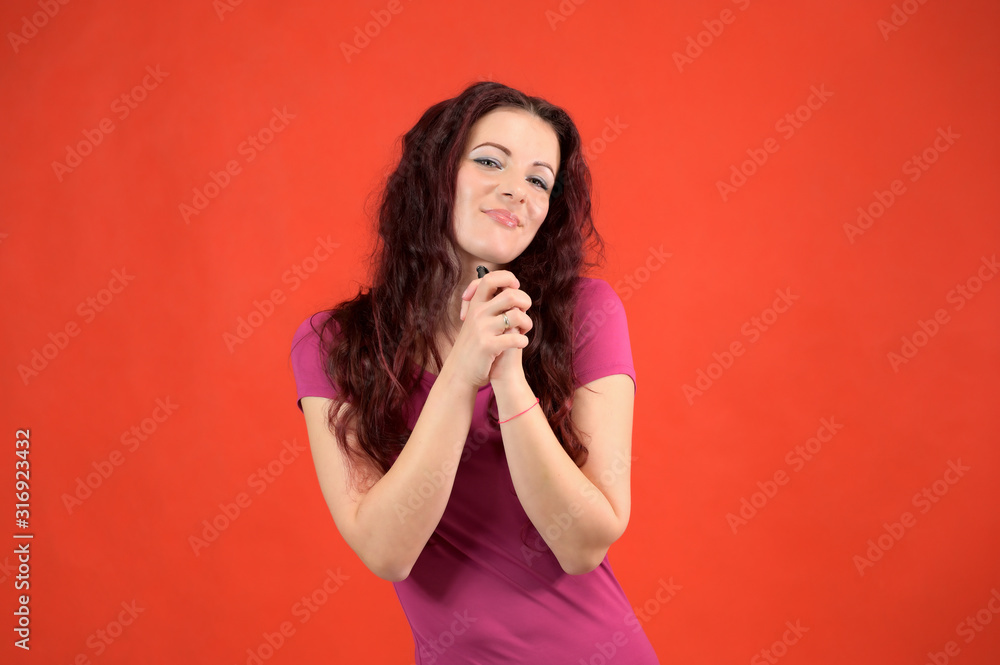 The model is smiling and talking cute to the camera. Portrait of a pretty slim brunette woman with emotions in a pink dress and with brown hair on a red background in the studio. The model is smiling and talking cute to the camera. Portrait of a pretty slim brunette woman with emotions in a pink dress and with brown hair on a red background in the studio.