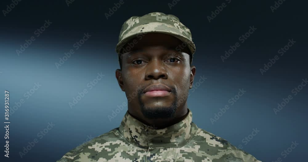 Close up of the sad serious face of the young African American man soldier in the uniform and hat looking straight at the camera on the dark background wall. Portrait. Close up of the sad serious face of the young African American man soldier in the uniform and hat looking straight at the camera on the dark background wall. Portrait.