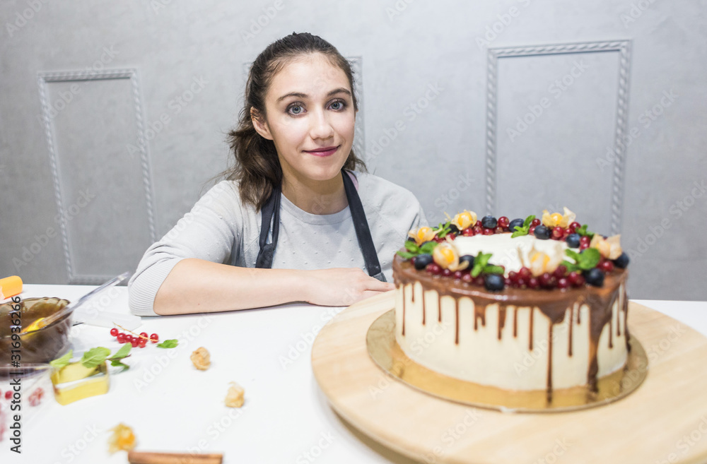 A confectioner stands next to a cooked biscuit cake with white cream, decorated with chocolate and berries. Cake stands on a wooden stand on a white table A confectioner stands next to a cooked biscuit cake with white cream, decorated with chocolate and berries. Cake stands on a wooden stand on a white table