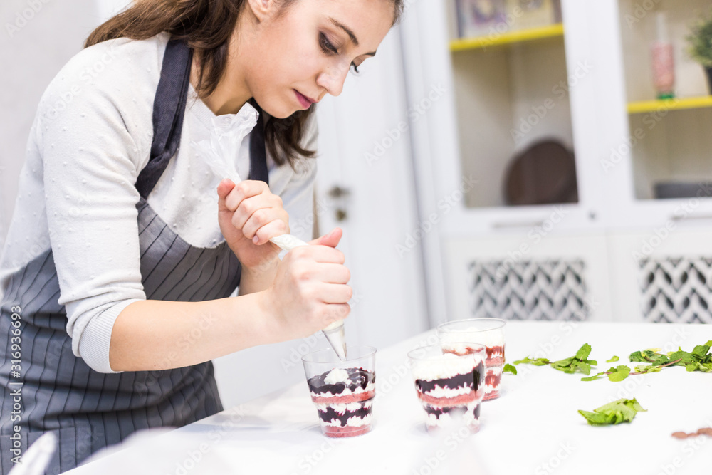 A confectioner prepares a trifle in three cups. Desserts are on the white table in the kitchen. The concept of homemade pastry, cooking cakes.