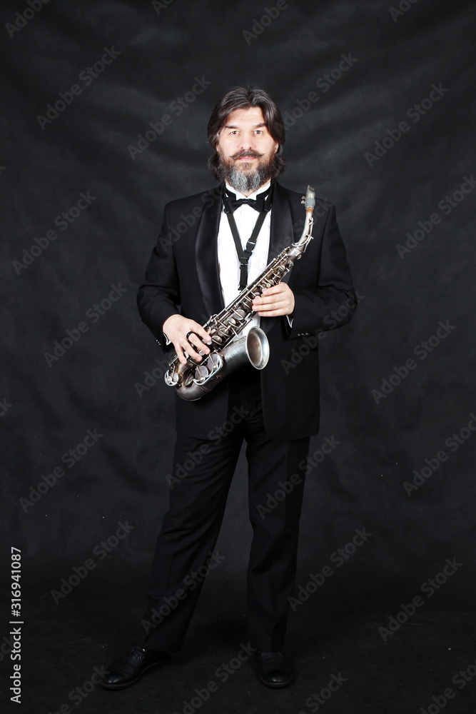 Fototapeta premium Pełne ujęcie profilowe przedstawiające muzyk artystę w klasycznym czarnym garniturze, we fraku, posągowe w muszce z brodą, gra muzykę na złotym saksofonie. Czarne tło