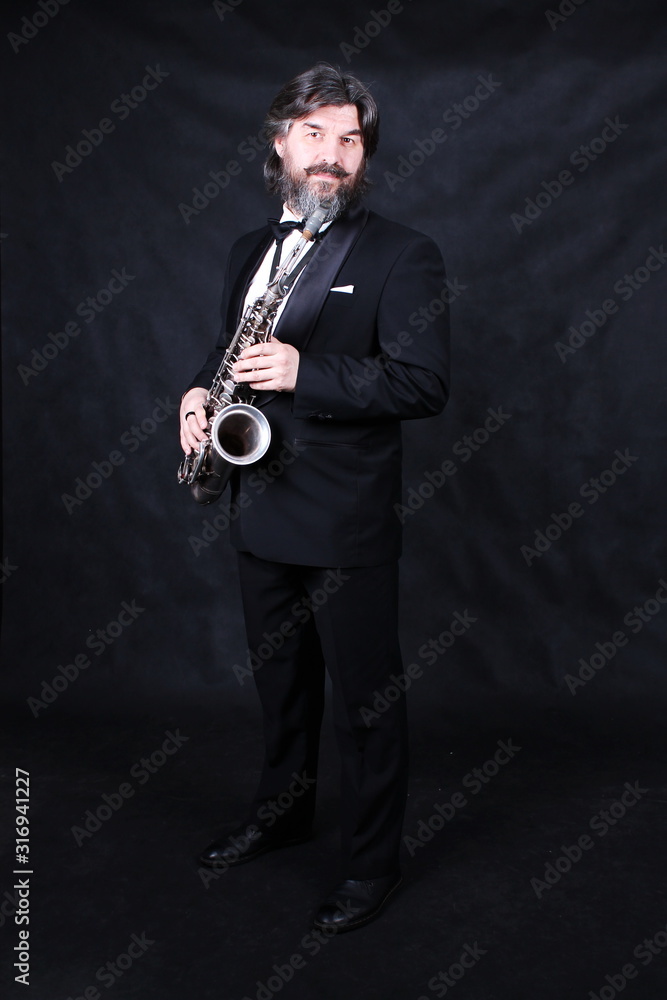 Naklejka premium Full length profile shot of male artist musician in a classic black suit, tailcoat, statuesque in a bow tie with a beard plays music on a gold saxophone.black background