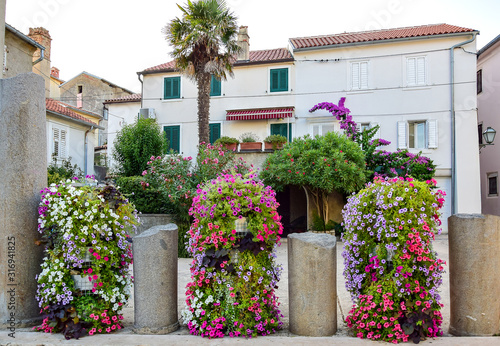 Krk, Croatia - Three large bulbs with colorful flowers between four dilapidated stone columns, a palm tree, trees with flowers and a white building with a brown roof in the background in the summer.