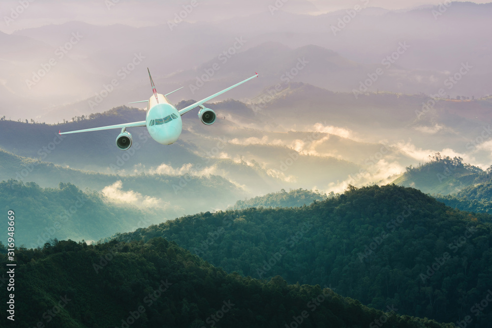 Fototapeta premium Airplane is flying over mountains in fog at sunset in winter. Landscape with passenger airplane, Business travel,Commercial plane,Aerial view.