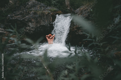 young red-haired girl bathes in a cold mountain waterfall in a green coniferous forest