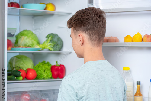 Back view - young teen boy standing near open fridge in kitchen at home. Portrait of pretty child choosing food in refrigerator full of healthy products - rear view.