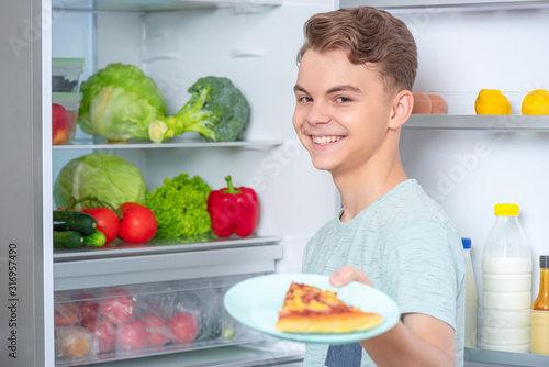 Hungry cute young teen boy holding plate with slice of pizza while standing near open fridge in kitchen at home. Portrait of pretty child choosing food in refrigerator full of healthy products.