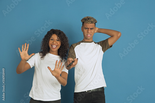 Young emotional african-american man and woman posing on blue background. Beautiful couple. Concept of human emotions, facial expession, relations, ad. Watch TV together, her favourite channel.