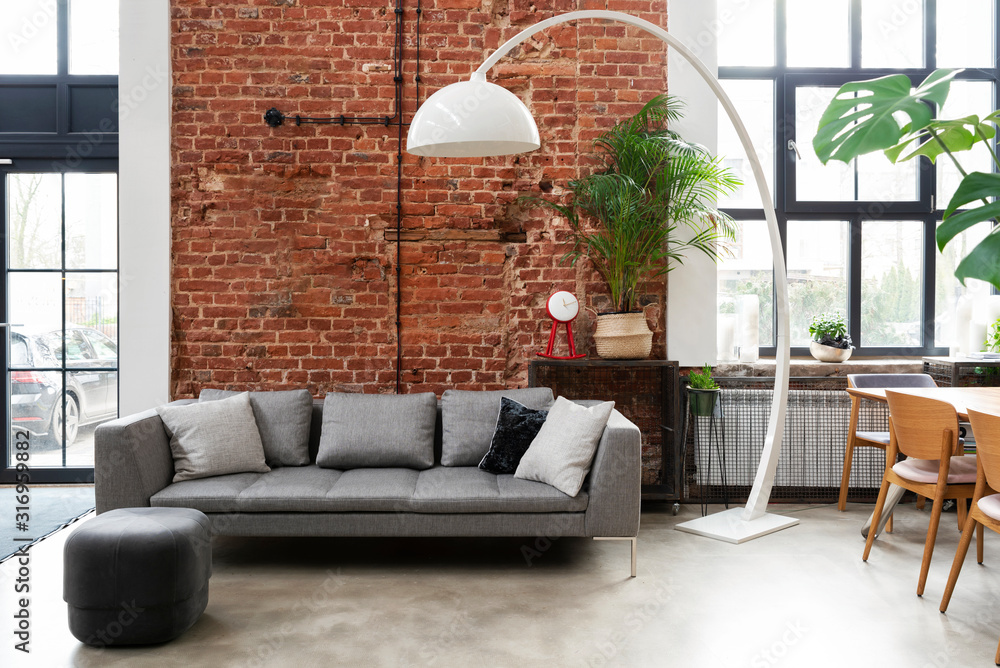 Living room interior in loft apartment in industrial style with brick wall, grey stylish sofa and big window. Modern lamp and design furniture in minimal indoors.  - obrazy, fototapety, plakaty 