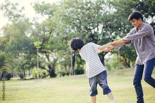 Happy Asian father playing and swinging his son in a park, Dad hold his kid hand to spin around on summer background