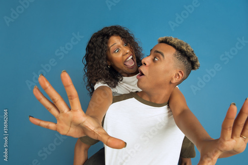 Young emotional african-american man and woman in white casual clothes posing on blue background. Beautiful couple. Concept of human emotions, facial expession, relations, ad. Hugging, laughting.