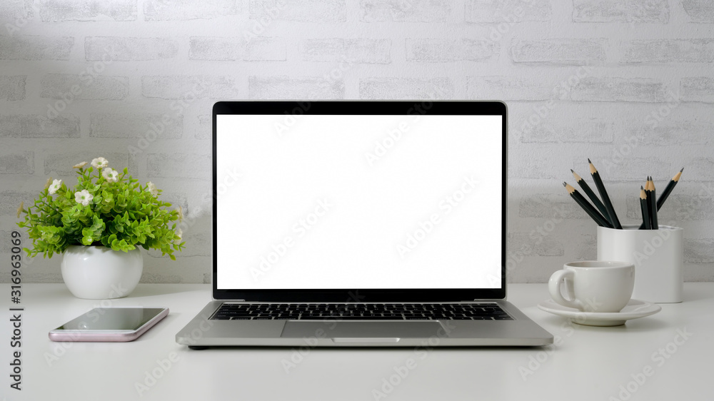 Close up view of workspace with blank screen laptop, smartphone, pencils, coffee cup and tree pot on white desk with brick wall Close up view of workspace with blank screen laptop, smartphone, pencils, coffee cup and tree pot on white desk with brick wall
