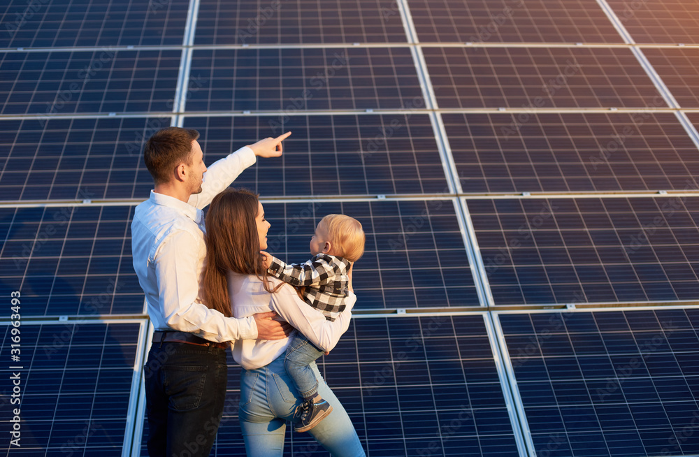 Husband shows his family the solar panels on the background. Blond child in arms of wife. Young family keeps up with the times choosing solar heating - obrazy, fototapety, plakaty 