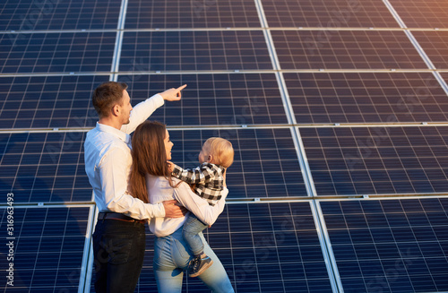 Husband shows his family the solar panels on the background. Blond child in arms of wife. Young family keeps up with the times choosing solar heating photo