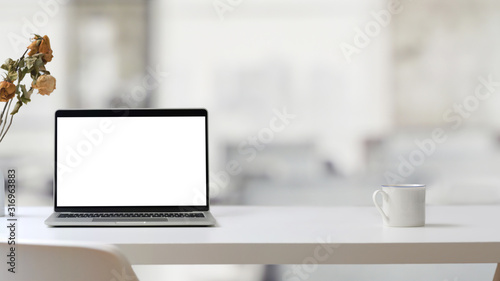 Close up view of workplace with open blank screen laptop and coffee cup on white table with blurred office room