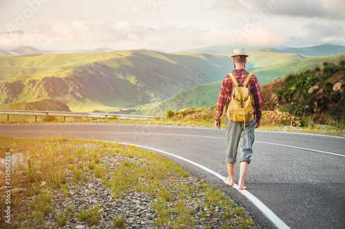 Fotografija Stylish barefoot bearded male hitchhiker traveler in a hat and with a backpack walks along a country road in the mountains at sunset