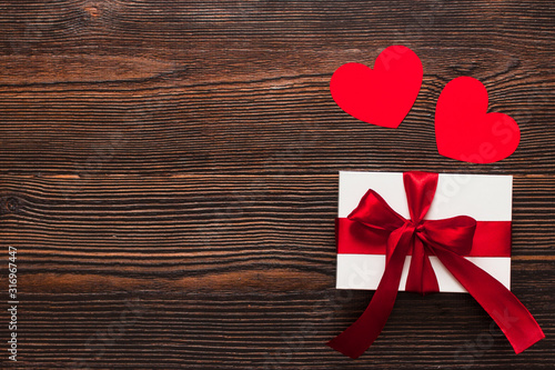 White present with a red ribbon and paper hearts isolated on a dark wooden background. Top side view of a celebrating warm flatlay. Valentines day and Christmas concept. Copyspace.
